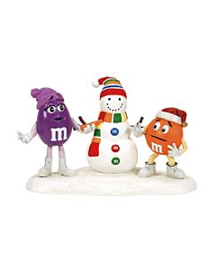 M&M’s characters Purple and Orange build a festive snowman; Purple playfully holds the carrot nose, teasing Orange in a lighthearted holiday moment