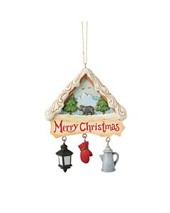 A cozy lodge ornament frames a bear wandering through a snowy forest with mountains in the background. Hanging charms like a lantern, canteen, and oven mitt add festive detail, while a “Merry Christmas” sign warms the scene.