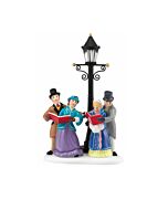 A group of villagers gathered beneath a glowing lamppost, singing Christmas carols on a snowy evening.