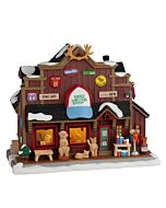 Charming miniature Wild Wooly's Gift Shop building with warm golden windows glowing against snowy Vail Village scenery, complete with detailed rustic architecture and cozy mountain lodge styling