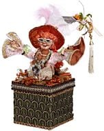 A whimsical collectible featuring an elaborately dressed red-haired witch, perched atop a decorative box adorned with colorful accents. The hand-crafted figure displays vibrant attire and a mischievous expression, embodying Mark Roberts' signature playful