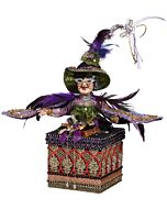 A lavishly adorned witch figurine, draped in regal purples and mossy greens, exudes opulence atop an intricately designed box. Her ensemble sparkles with sequins, jewels, and feathers, capturing the essence of luxury and enchantment.